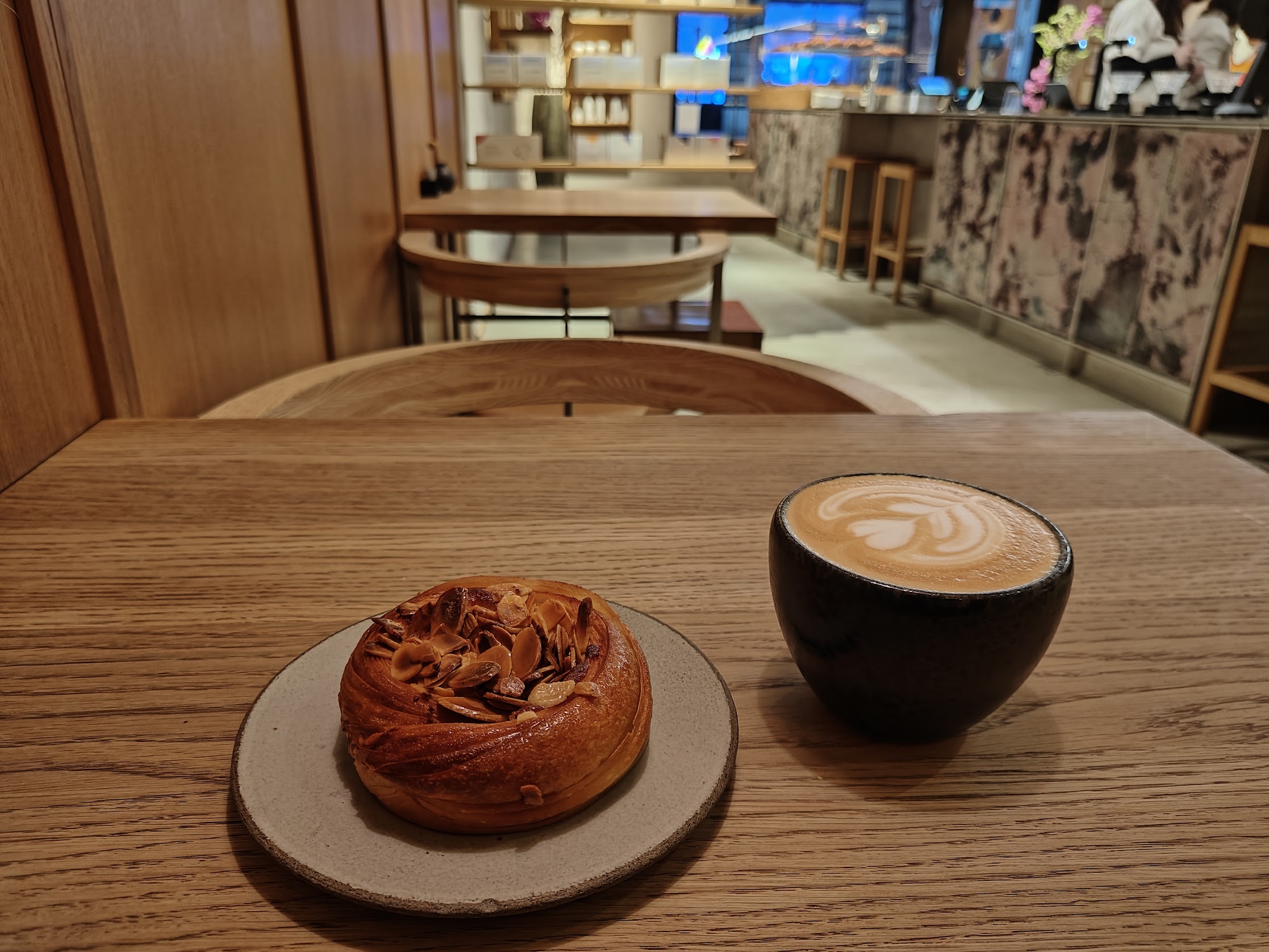 A latte with heart-shaped foam art sits in a dark ceramic cup beside a flaky almond pastry on a small plate, placed on a wooden table inside a warm, modern café with shelves, stools, and the counter softly blurred in the background